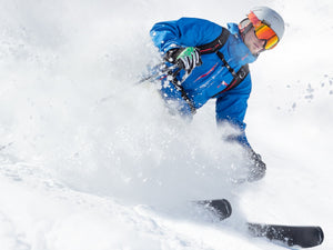 Man in a blue jacket, white helmet and orange snow goggles, skiing down a snow slope with a large snow cloud